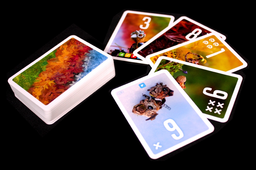 A deck of colorful playing cards with a vibrant autumn-themed card on the top, surrounded by cards displaying whimsical characters and numbers.