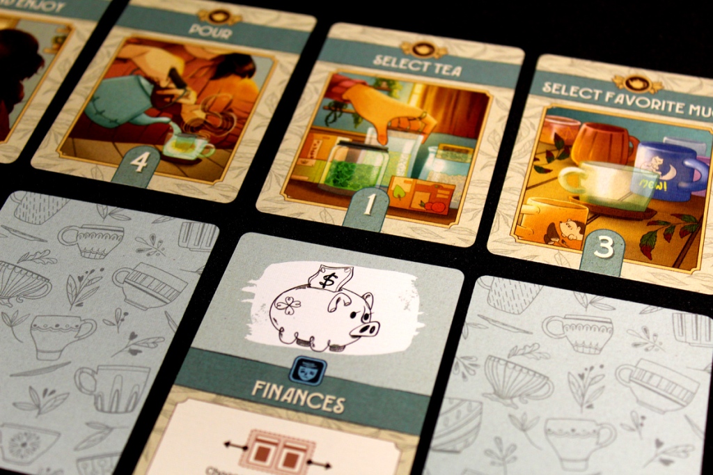 Close-up of cards from the game A Nice Cuppa, featuring actions like 'Select Tea,' 'Pour,' and 'Select Favorite Mug.' A piggy bank illustration labeled 'Finances' is also visible.